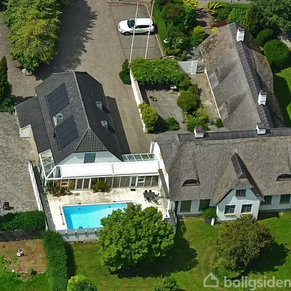 En stor ejendom med to huse omgivet af træer. Det ene hus har et stråtækt tag, mens det andet har solpaneler. En swimmingpool ligger i gården mellem dem. Tekst: "boligsiden".