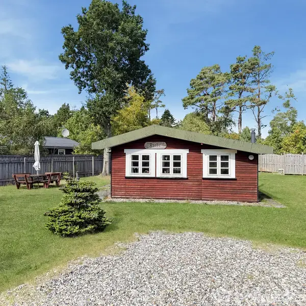 Rødt træhus med grønne skodder står omgivet af en græsplæne og have med træer og stakit. Huset har et skilt med teksten "Solhøjen". En havebordsgruppe ses til venstre.