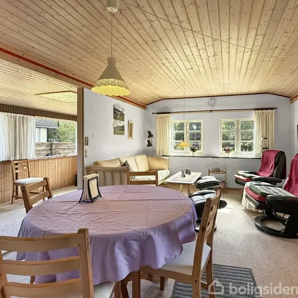 Rundt spisebord med lilla dug står i en lys stue med træloft og store vinduer. Komfortable stole og en sofa står ved siden af et sofabord, omgivet af træpanelvægge.