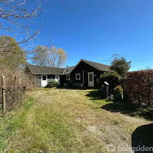 Et sort træhus står stille på en græsbeklædt grund med en garage ved siden af. Flere træer omkranser huset, og en bil er parkeret til højre. Nummer "33" ses ved indgangen.