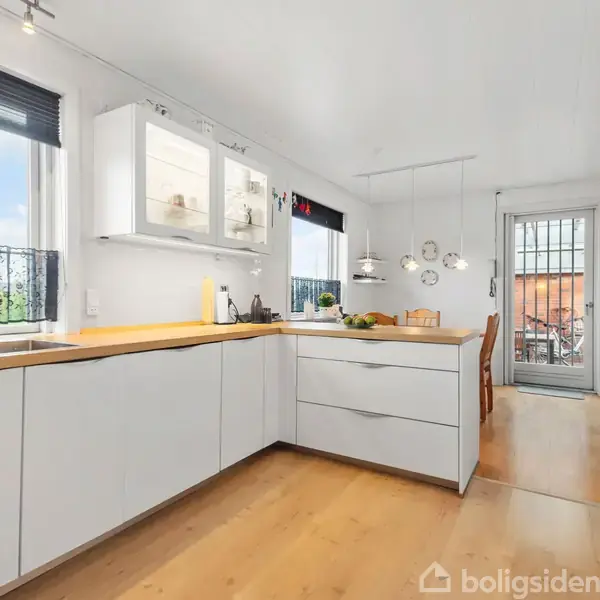 Køkken med hvide skabe og træbordplader. En vask står ved et vindue med persienner. Lamper hænger over en centralt placeret køkkenø. Gennem en dør ses en gårdhave.