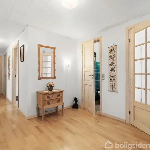 Gang med lyst trægulv har et bord med blomster og spejl på væggen. Døre fører til forskellige rum; en viser glimt af et badeværelse. Vægpynt fuldender indretningen.