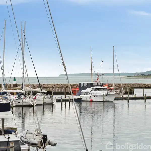 Både ligger stille ved en havn med stille vand, omgivet af en mole og en blå himmel i baggrunden.