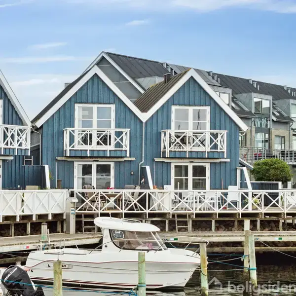 Blå træhuse står stille ved en bådehavn, med både fortøjede til pæle i forgrunden. Træhytterne har hvide altaner, og baggrunden viser en ren og klar himmel.