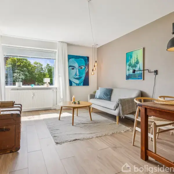 Stue med sofa, sofabord og spisebord. Tv hænger på væggen. Store vinduer slipper naturligt lys ind. Dekorativt kunstværk på væggen, trægulv, grønne omgivelser udenfor.