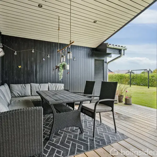 Overdækket terrasse med sortmalede trævægge og loft, indeholder grå hjørnesofa, sort spisebord og stole. Terrasse omgivet af grøn græsplæne og tæt hæk i baggrunden.