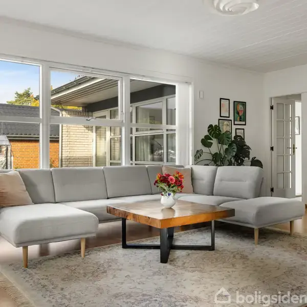 Grå hjørnesofa står i lys stue med store vinduer. Der er et træbord med en vase med blomster foran sofaen. Billeder hænger på væggen, og planter står i hjørnet.