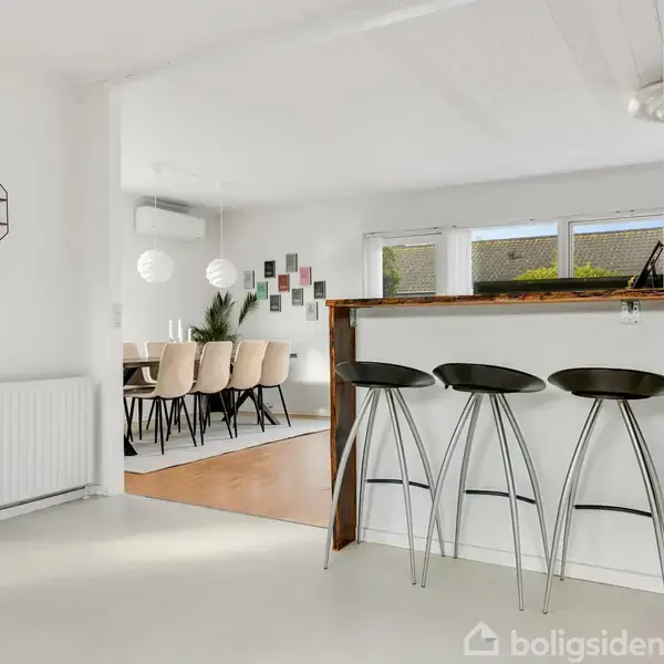 Tre sorte barstole står ved en træbar; i baggrunden ses en spisestue med beige stole og et bord med hængende lamper. Vægdekorationer og store vinduer giver lys.