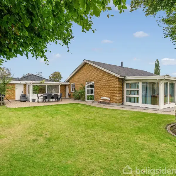 Et gulstenshus står i en grøn have med en velplejet græsplæne. Til venstre er en terrasse med havemøbler og grill, omkranset af buske og træer under blå himmel.