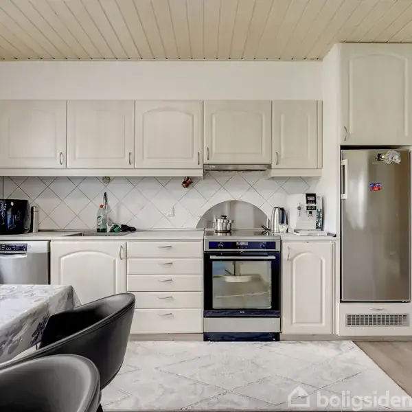 Et lyst køkken med hvide skabe og apparater, herunder en ovn, opvaskemaskine og køleskab. Et bord med dug og stole står ved siden af, flasker og kaffemaskine på bordet. En ur hænger på væggen.