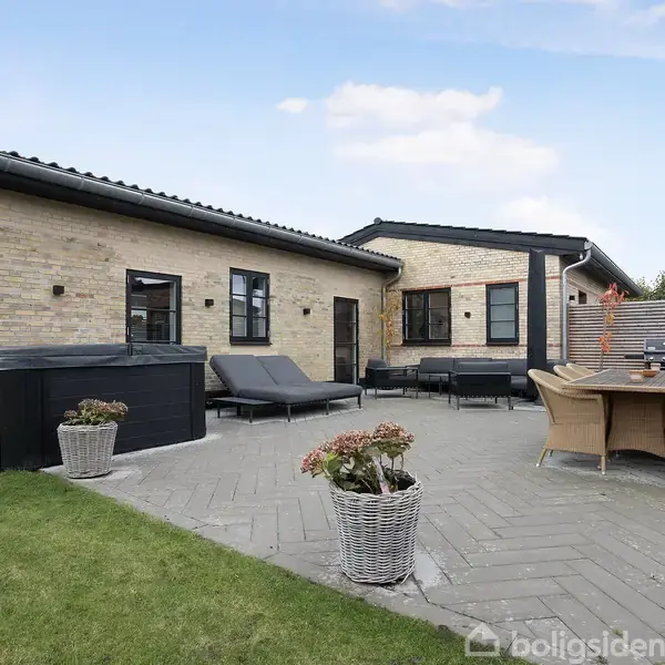 Hus i mursten, med fladt tag, omgivet af fliser og en grøn græsplæne. Terrassen har loungemøbler, et bord med stole og blomsterkrukker.