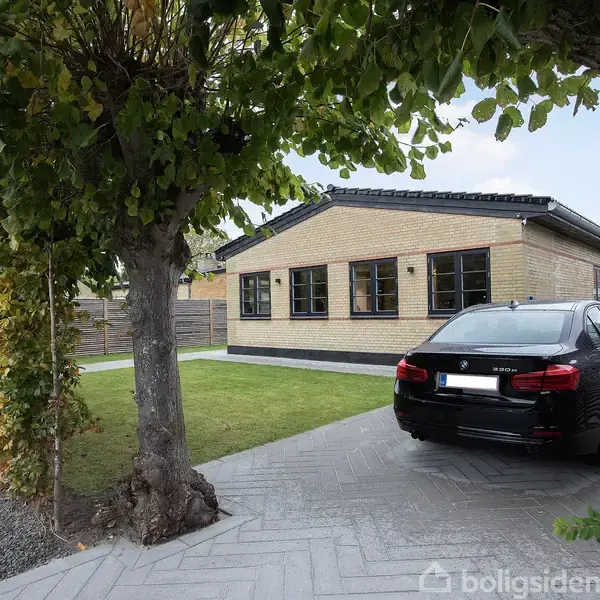 En sort bil holder parkeret på en indkørsel foran et moderne, ens-etages hus med gule mursten. Omgivet af grønne træer og velplejet hæk i et forstadskvarter.