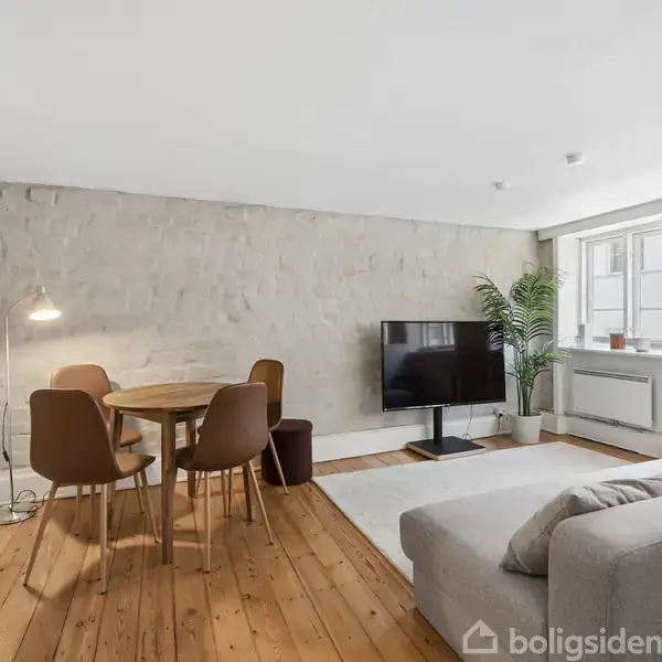 En hyggelig stue har en grå sofa foran et mørkt TV, mens et rundt bord med tre stole står tæt ved en væg. Trægulve og store vinduer giver naturligt lys.