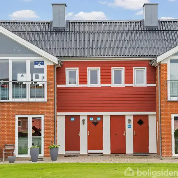 Rødt rækkehus med store glaspartier og tre døre, omgivet af en græsplæne foran. To altaner på hver side over døre. Blå skilt med "Til leje" hænger i venstre vindue.