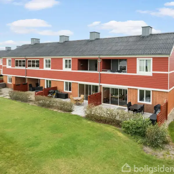 Rækkehuse i rød træbeklædning står stille ved en grøn græsplæne. Hver enhed har egen terrasse. Skyfri blå himmel med hvide skyer giver en klar og åben atmosfære.