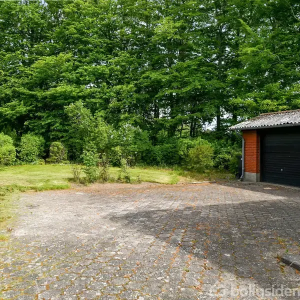 Garagebygning står stille langs en brolagt indkørsel omgivet af grønne buske og en skovgrænse i baggrunden. Indkørslen er rummelig med en græskant.