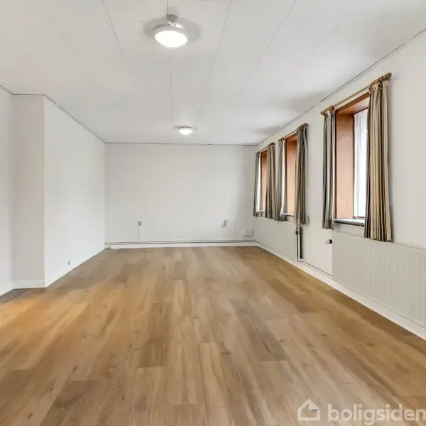 En rummelig, tom stue med brune trægulve, vinduer med stribede gardiner og hvide vægge. Loftsbelysning er tændt, og der er en radiator langs væggen.