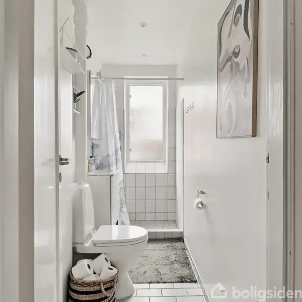 Et smalt badeværelse med et toilet i forgrunden. Et bruseområde ses ved vinduet bagved. Hvide vægge og gulve omgiver rummet. En kurv står ved siden af toilettet.