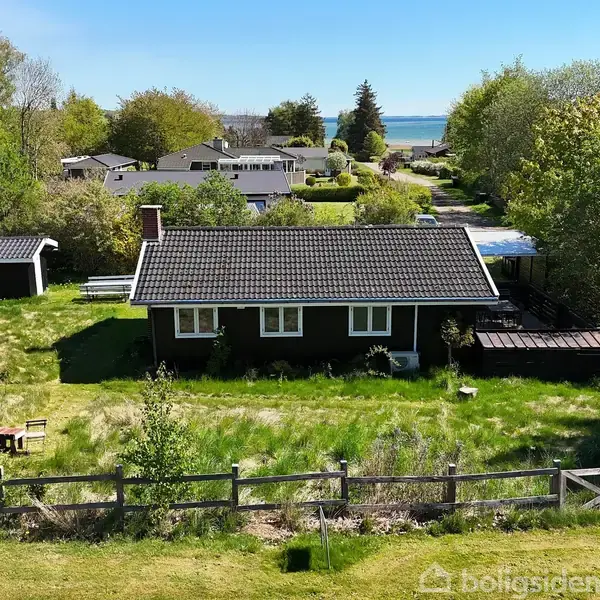 Hus står stille i grøn have med græs og træer, omgivet af træer og buske. Flere huse i baggrunden, med udsigt til blå horisont og hav.