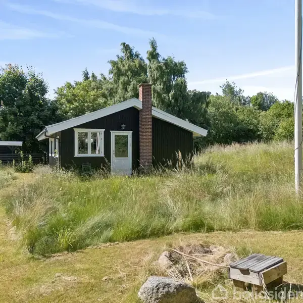 Et lille, sort-hvidt sommerhus med skorsten står midt på en overgroet græsplæne. Til venstre ses et bord med stole. I baggrunden er der frodig vegetation.