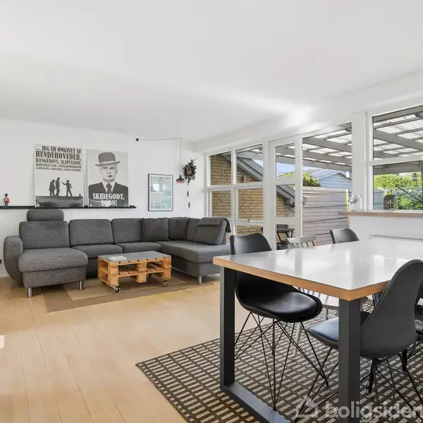 Stue med grå hjørnesofa og træsofabord, placeret ved siden af spisebord med sorte stole. Store vinduer giver udsigt til terrasse og have. Plakat på væggen viser mand i hat.