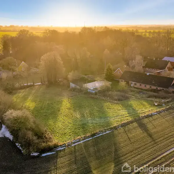 En landbrugsejendom ligger stille under en klar himmel med solstråler, der kaster lange skygger på de omkringliggende marker. Træer og bygninger omkranser stedet.