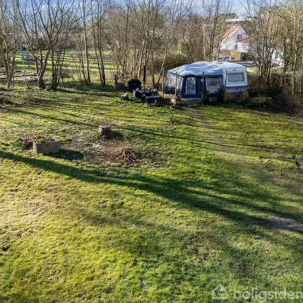 Campingvogn står stille på en grøn, græsbeklædt grund omgivet af nøgne træer. Et bord og et par stole er placeret udenfor. I baggrunden ses en bygning.