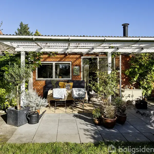 Overdækket terrasse med potteplanter og møbler foran et rødt murstenshus. Solrigt vejr og en velholdt have omkranser området.
