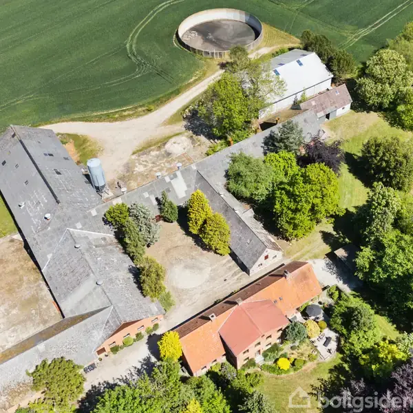 Gårdkompleks med flere bygninger omgivet af grønne marker og træer, set fra luften. En rund silo ses i øverste kant omgivet af grusveje.