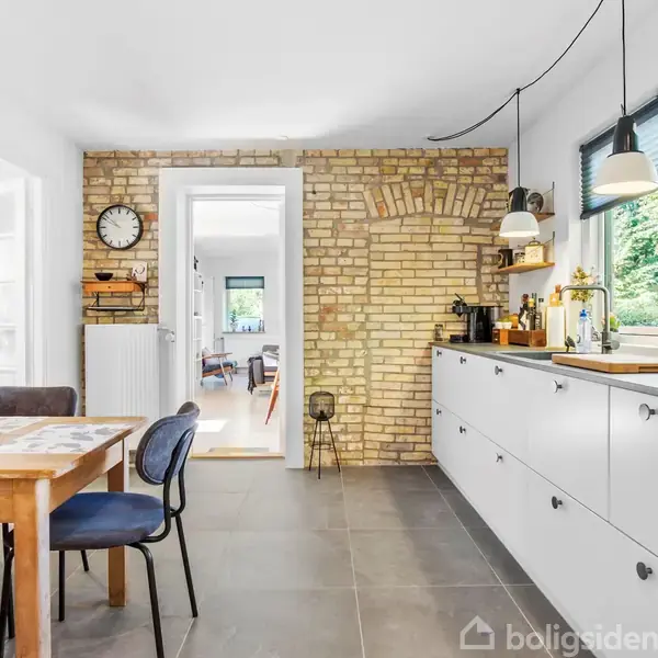 Køkkenbord med stole ved siden af et moderne køkken med hvide skabe. En murstensvæg med en stor ur hænger over radiatoren. Store vinduer giver udsigt til grønne omgivelser.