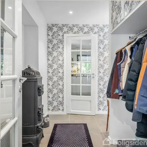 En gang med en gammel brændeovn til venstre, flere jakker hængende til højre, og en glasdør med blomstret tapet i midten, fører til et lysere rum.