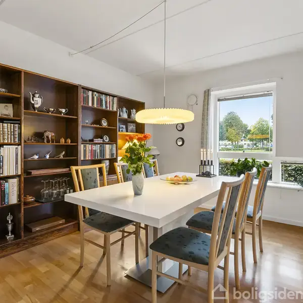 Et spisebord med fire stole står i et lyst rum. En reol fyldt med bøger og pynt genstande ses bagved. Et vindue giver udsigt til træer udenfor.