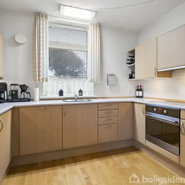 Et moderne køkken viser lyse træskabe og grå bordplader. Køkkenmaskiner står på venstre side. Et vindue med persienner lukker lys ind. En ovn er indbygget til højre.