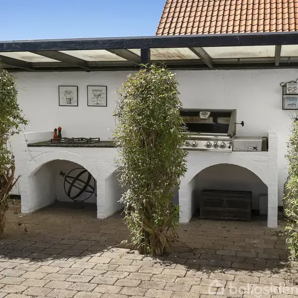Udendørs køkken med gasgrill og kogeplade under halvtag. Hvid murstensvæg prydet med små rammer. Omgivet af grønne buske og flisebelagt område.