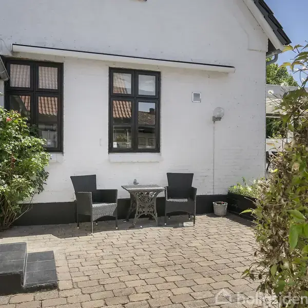 To sorte stole og et rundt metalbord står på en flisebelagt terrasse foran en hvid murstensbygning med sorte vinduer. Planter omkranser området.