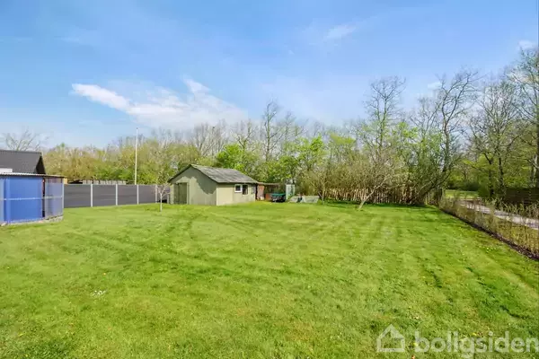 En græsplæne strækker sig foran en lille, grøn bygning. Omkring bygningen står høje træer og et gråt hegn. Himlen er blå med spredte skyer.