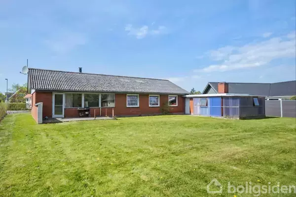 En murstensbygning med sort tag står stille i en grøn have med græsplæne og et tilstødende skur til højre. Huset har store vinduer og en satellitmodtager.