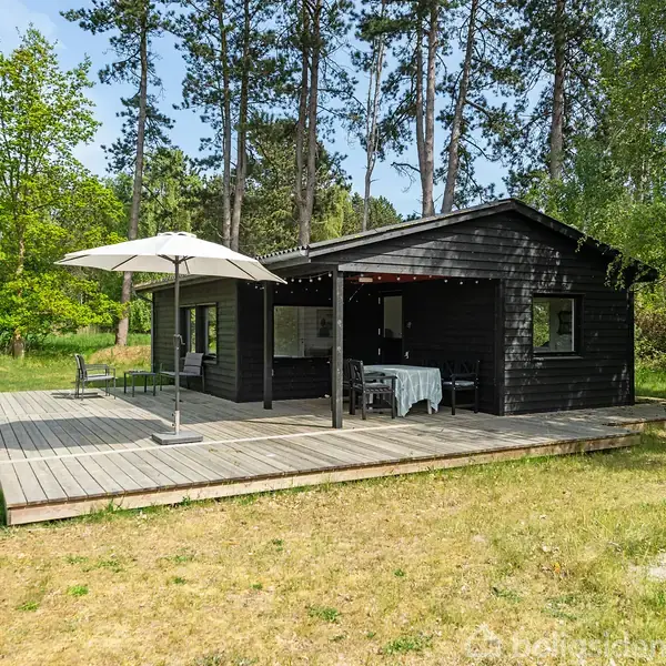 En sort, træsommerhus med åbne glasdøre står på en terrasse med en parasol og havemøbler. Omgivet af høje træer og græs i en skovklædt område.