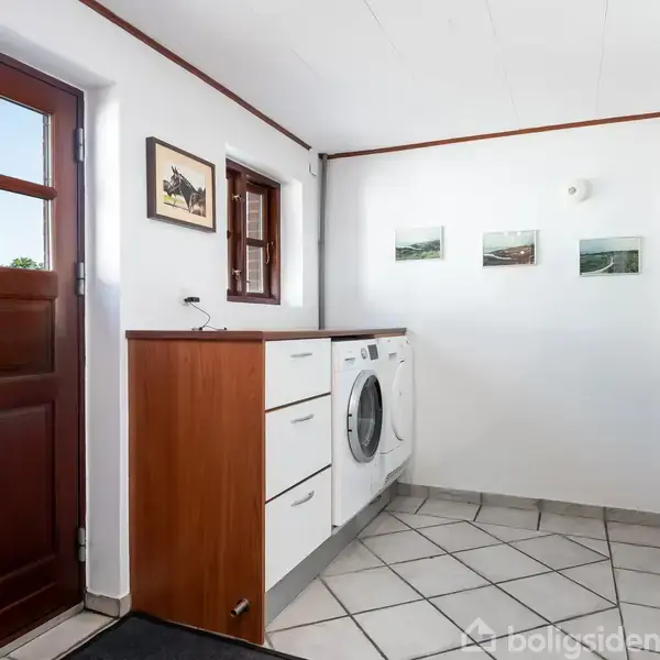 Vaskemaskine står ved siden af hvidt skab i rum med flisegulv. Trædør til venstre, væg med tre små billeder. Rundt spejl og hvid væg i baggrunden.