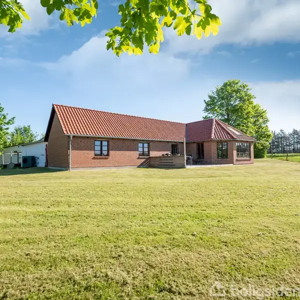 En rød murstensvilla med rødt tegltag står på en stor grøn græsplæne, omgivet af træer og et åbent landskab under en blå himmel.