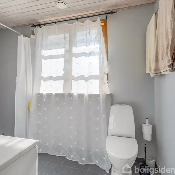 Toilet står stille ved siden af en holder med toiletpapir i et lyst badeværelse med gulvfliser og et vindue med hvidt gardin. Håndklæder hænger på væggen.