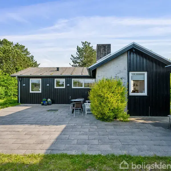 Et sortmalet hus med terrasse, hvor stole og et bord står. Omgivet af grønt græs og træer under en blå himmel.