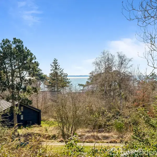 Et mørkt træhus står stille omringet af træer og buske, med en vej foran. I baggrunden anes en rolig udsigt til havet under en klar blå himmel.