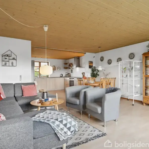 En stue med gråt sofahjørne og to stole omkring et kaffebord. I baggrunden ses et åbent køkken og spisebord med stole. Væggene har dekorative tallerkener, og der er to vitrineskabe.