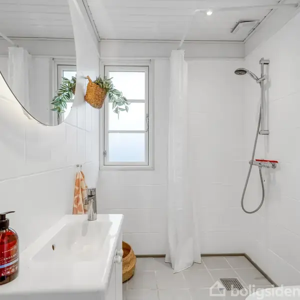 Badeværelse med brusebad til højre, hvide klinker, spejl over vasken med sæbedispenser og planteophæng. To vinduer slipper lys ind. Gardin adskiller bruseren fra resten af rummet.