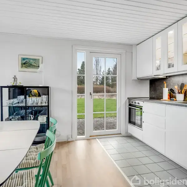 Moderne køkken med hvide skabe, hvor spisebordet har grønne stole. En glasdør fører ud til en have. Friske blomster og køkkenredskaber er på bordet og ved komfuret.