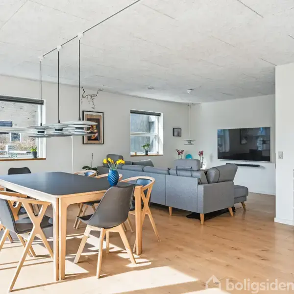 Spisebord med stole, arrangeret under moderne lamper, står i et lyst rum. Bagved ses en stue med sofagruppe, tv på væggen, og et skab til højre. Vaser med blomster pynter rummet.