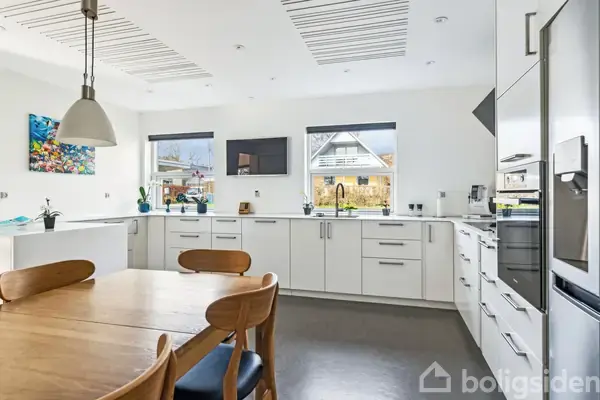 Køkkenbord står i fokus i moderne køkken. Det er omgivet af hvide skabe og apparater med trægulve og store vinduer, der lader lys ind. Maleri hænger på væggen.