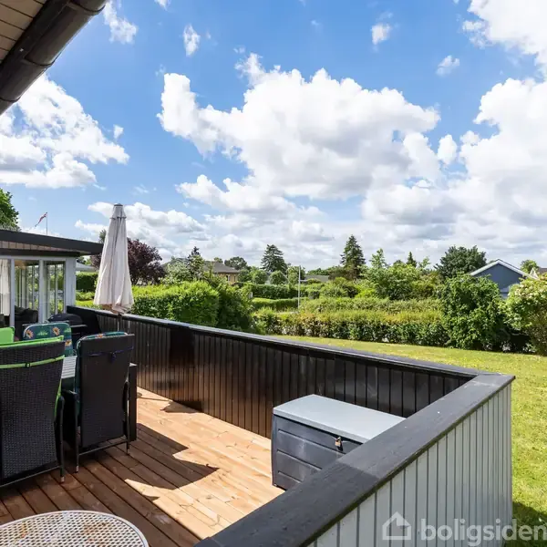 Terrassemøbler står stille på en træterrasse med udsigt over en grøn have. En hæk adskiller terrassen fra en græsplæne. Skyer bevæger sig langsomt på den klare himmel.