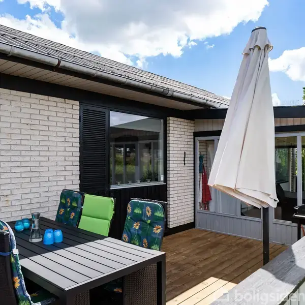 Terrasse med spisebord og fire stole med blomstrede hynder; der står også to blå lysestager på bordet. En stor parasol er opstillet, omkranset af hvid murstensvæg og grønne træer.